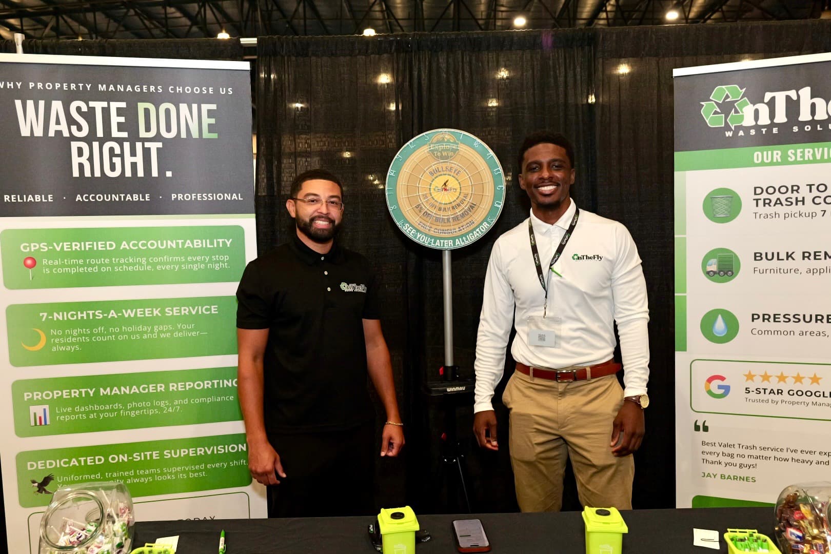 Two On The Fly Waste Solutions team members standing at the company booth, with branded banners and a prize wheel, at a Central Florida property management trade show